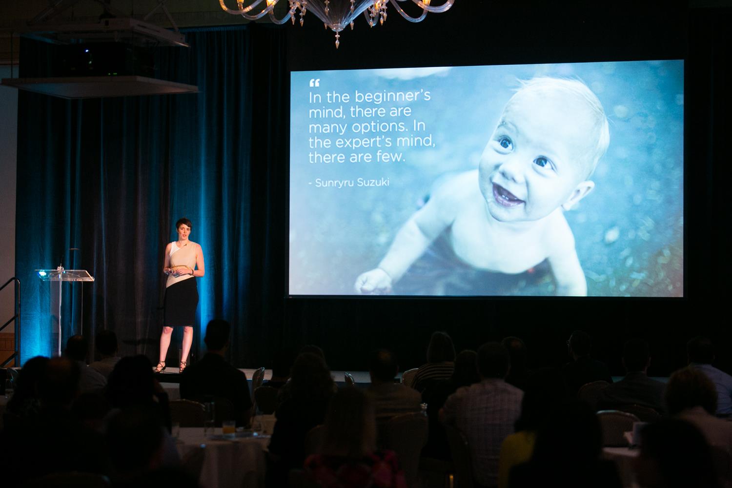 Brooks Bell speaking on stage at a conference, large chandelier and audience visible
