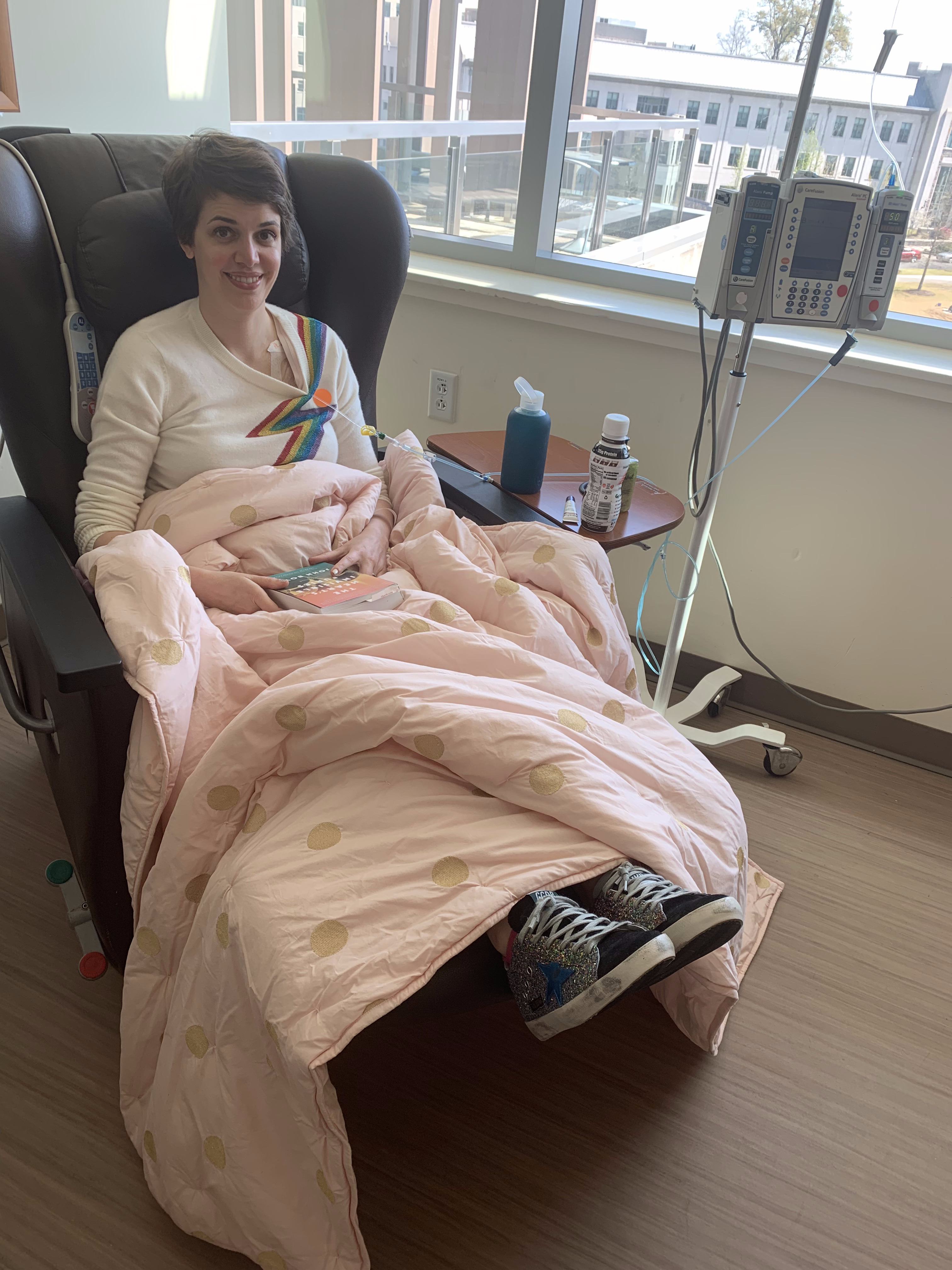 Brooks Bell in the chemo chair, smiling with a rainbow lightning bolt sweater and pink polka dot blanket