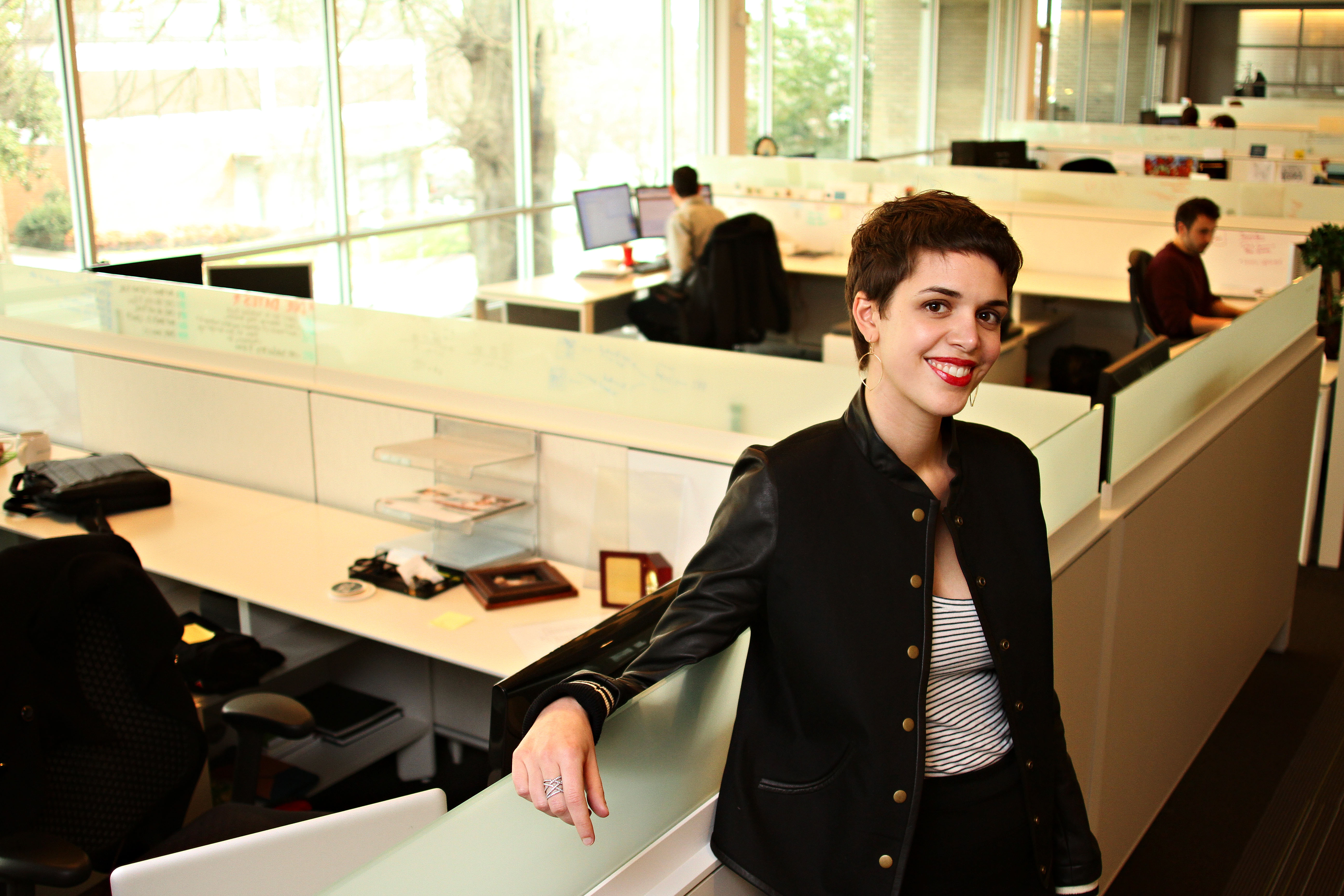 Brooks Bell leaning on a desk in her office, team members working behind her
