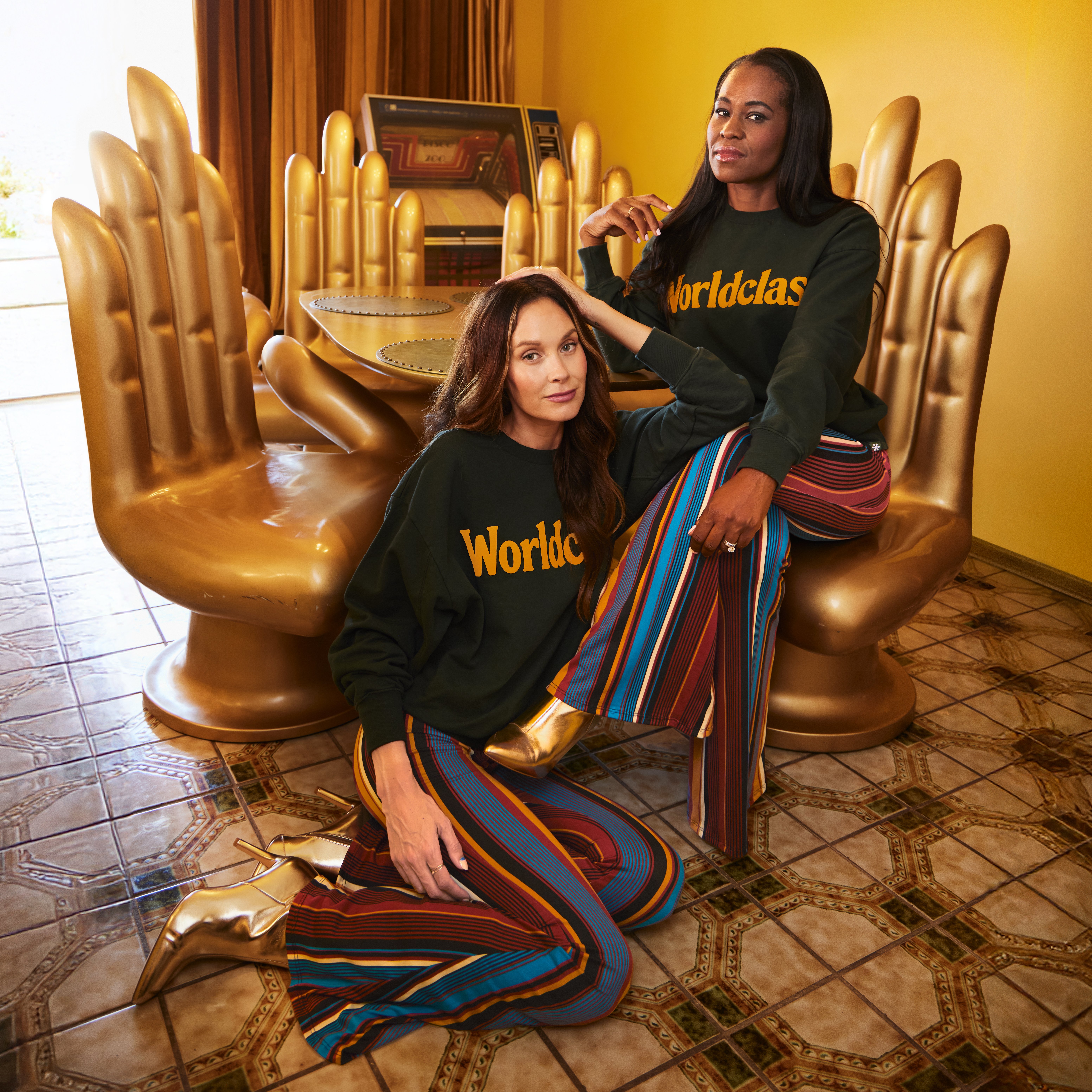 Two women wearing Worldclass sweatshirts in an editorial setting with gold hand chairs
